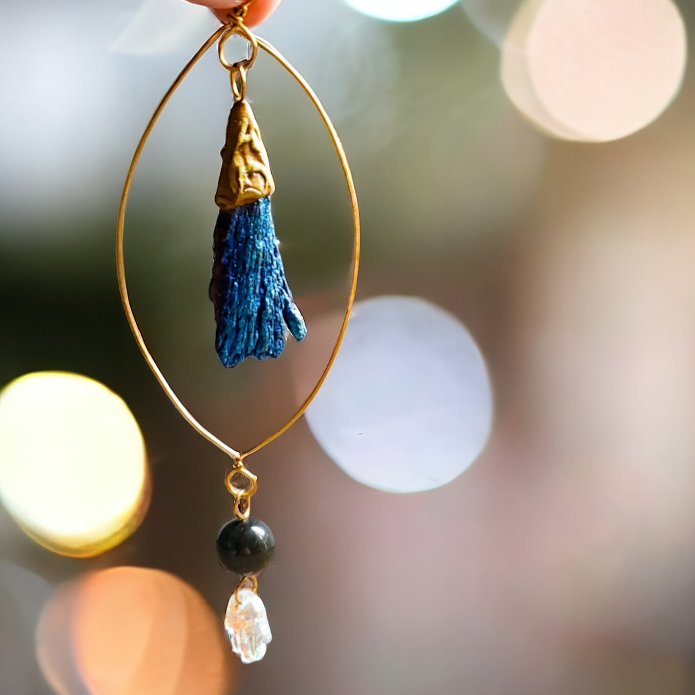 Crystal Suncatcher with hammered wire eye design, featuring an aura-coated Black Kyanite fan set in detailed brown polymer clay. Dangling elements include a Gold Sheen Obsidian bead and a glass crystal Hamsa Hand. Approximately 5.5 inches long, designed for protection and witchy decor near the front door. Powerful symbolism with crystals for protection and enchanting aesthetics.
